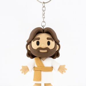 May include: A keychain featuring a cartoon-style figure of Jesus. The figure has brown hair and a beard, wearing a white robe with a gold sash. The keychain includes a silver-colored ring and chain.