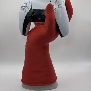 May include: A red hand-shaped controller stand holding a white and black video game controller. The stand has a gray base. The controller has buttons and directional pads.