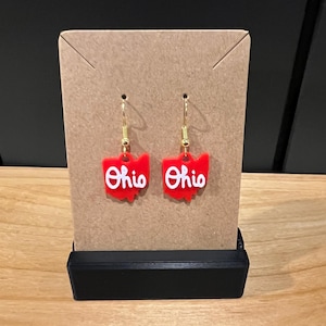 May include: Red Ohio state-shaped earrings with gold hooks and white lettering. The earrings are displayed on a brown card with a black base.