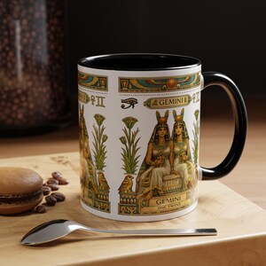 Puede incluir: Taza de cerámica blanca con interior y asa negros. La taza presenta un diseño de temática egipcia con ilustraciones de figuras, jeroglíficos y el texto "GEMINI THE TWINS" en dorado y turquesa.