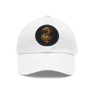 Puede incluir: Gorra de béisbol blanca con un parche circular que muestra una ilustración detallada de la cabeza de un águila dorada. El águila lleva un collar dorado sobre un fondo negro. La gorra tiene una visera curva y un diseño clásico ajustable.