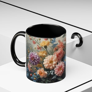 Puede incluir: Una taza de café con asa negra y diseño floral. La taza presenta un vibrante ramo de flores en tonos rosas, amarillos y morados sobre un fondo apagado. El interior de la taza es negro.