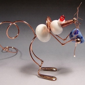 Pode incluir: Uma escultura de rato caprichosa de arame segurando uma conta com um padrão azul e branco. O rato é feito de arame de cobre e tem uma conta branca para a cabeça, com contas vermelhas para os olhos. A cauda é enrolada em espiral.