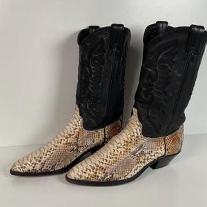 May include: A pair of mid-calf cowboy boots featuring black leather shafts with decorative stitching and pointed toes. The lower portion of the boots is constructed from a textured, snakeskin-patterned material in shades of beige, tan, and brown.