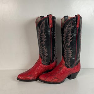 May include: A pair of cowboy boots with a red snakeskin pattern on the foot and a black shaft. The shaft features decorative stitching in a lighter red color, with a red trim. The boots have a pointed toe and a black heel.
