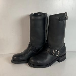 Puede incluir: Un par de botas de moto de cuero negro. Las botas son de media caña, con punta redonda y tacón bajo. Cada bota tiene una correa con hebilla alrededor del tobillo y un acabado liso y pulido. Las botas están diseñadas para la durabilidad y el estilo.