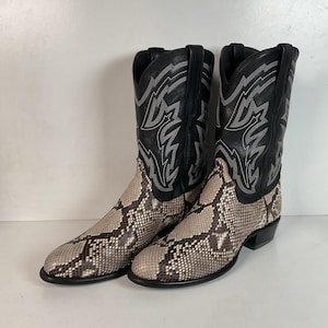 May include: A pair of cowboy boots with a snakeskin pattern on the lower part and black leather uppers. The black leather uppers have decorative stitching in a silver color. The boots have a pointed toe and a stacked heel.