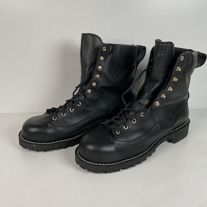 May include: Pair of black leather work boots with gold-colored eyelets and laces. The boots have a thick, lugged sole with white stitching. The upper part of the boots is tall, reaching up the ankle. The boots are displayed on a white surface.