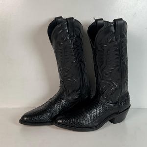 May include: A pair of black cowboy boots featuring a textured snakeskin-patterned vamp and a smooth leather shaft with intricate tonal stitching. These boots have a pointed toe, a stacked heel, and pull tabs at the top of the tall, structured shafts.