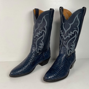 May include: A pair of navy blue cowboy boots featuring a textured, scale-like pattern on the foot and a smooth leather shaft. The upper portion of the boots displays intricate light blue decorative stitching and pull tabs at the top edges.