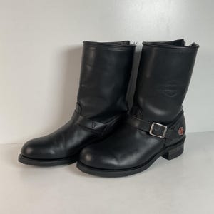 Puede incluir: Par de botas de moto de cuero negro con una correa con hebilla y un pequeño logotipo en el lateral. Las botas tienen una punta redonda y un tacón corto. El cuero parece ligeramente desgastado, dándoles un aspecto vintage.