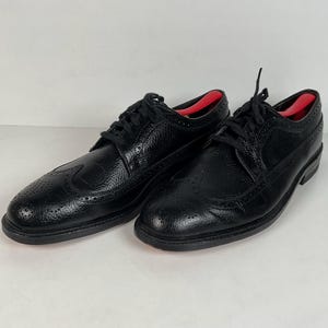 May include: A pair of black leather dress shoes with a textured finish. The shoes feature a wingtip design with decorative perforations and black laces. Red shoe trees are visible inside the shoes.