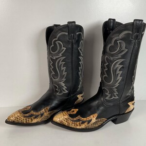 May include: A pair of black leather cowboy boots featuring a pointed toe with a tan snakeskin-textured overlay. The tall shafts are decorated with intricate grey stitching in a swirling pattern, and the boots have a stacked heel and pull tabs.