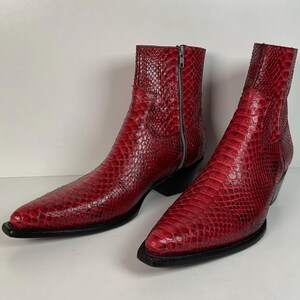 May include: A pair of ankle boots featuring a vibrant red snakeskin texture, a pointed toe, a stacked black heel, and a silver-toned side zipper closure. The boots have a structured silhouette with a clean finish and a black sole.