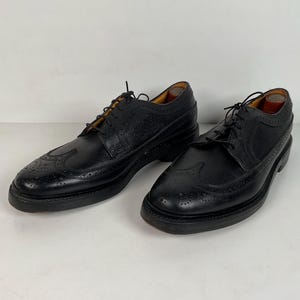 May include: A pair of black leather dress shoes with brogue detailing and a textured pattern on the upper. The shoes have a classic lace-up design and a thick sole. The shoes are displayed on a white surface.