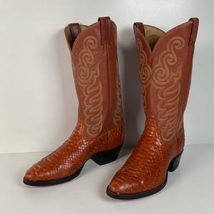 May include: A pair of cognac-colored leather cowboy boots featuring a textured, scale-like pattern on the foot and a smooth shaft with intricate, light-colored decorative stitching. The boots have a pointed toe, a stacked heel, and pull tabs.