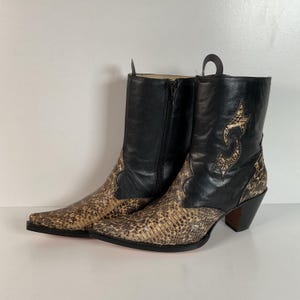 May include: A pair of cowboy boots with a pointed toe. The boots feature a black leather upper with a decorative design and a snakeskin-patterned lower section. The boots have a zipper closure and a stacked heel.