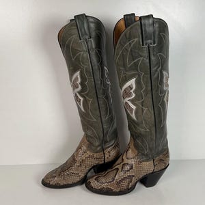 May include: A pair of knee-high western boots featuring a grey leather shaft with white stitched butterfly detailing and a textured snakeskin-patterned leather foot. The boots have a pointed toe, a stacked block heel, and pull tabs at the top.