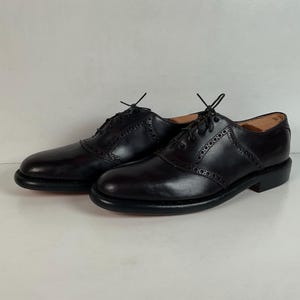 May include: A pair of black leather saddle shoes with a classic design. The shoes feature a rounded toe, lace-up closure, and decorative perforations. The soles are dark, and the shoes are set against a plain background.