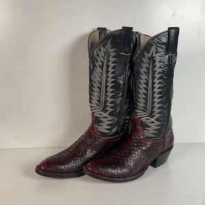May include: A pair of cowboy boots with a black upper and a red, textured lower section. The boots have decorative stitching in a light color and a pointed toe. The heels are black.