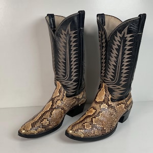 May include: A pair of tall cowboy boots featuring a textured snakeskin pattern on the foot and toe area, contrasted with black leather shafts. The black shafts are decorated with intricate tan stitching in a jagged, flame-like design.