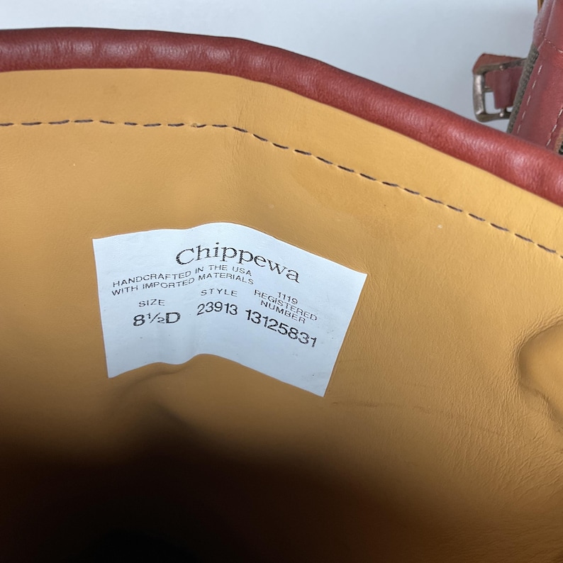Może przedstawiać: Wnętrze brązowego sk&oacute;rzanego buta z beżową wyści&oacute;łką i białą metką. Na metce widnieje napis Chippewa, Handcrafted in the USA with imported materials, rozmiar 8.5D, styl 23913 oraz numer rejestracyjny 1119 13125831. Widoczna jest metalowa klamra.