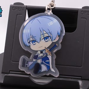 Puede incluir: Un llavero acrílico transparente con un personaje de anime chibi de pelo y ojos azules, sosteniendo una espada azul. El personaje viste ropa blanca y azul. El llavero está sujeto a un anillo de metal plateado.