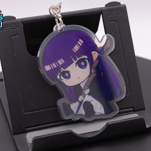 Puede incluir: Un llavero de anime de color morado y blanco con un anillo plateado. El personaje tiene el pelo largo morado y sostiene una espada. El llavero está sobre un fondo negro.