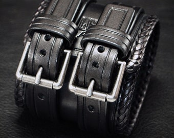 Op de afbeelding: Een zwarte leren armband met twee zilveren gespen en een gevlochten zwarte leren band.