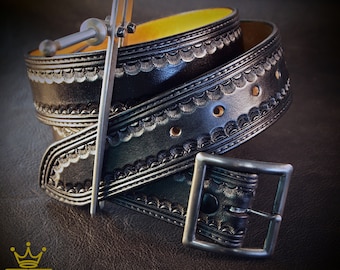 Puede incluir: Un cinturón de cuero negro con un diseño en relieve intrincado y una hebilla plateada. El cinturón está doblado por la mitad y la hebilla es visible.