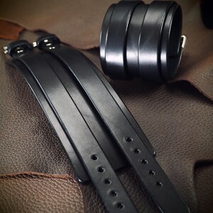 May include: Black leather wrist cuffs and matching bracelets. The cuffs are wide, with multiple stacked bands and a silver buckle. The bracelets have adjustable straps with multiple holes and silver hardware. The items are displayed on a textured brown surface.