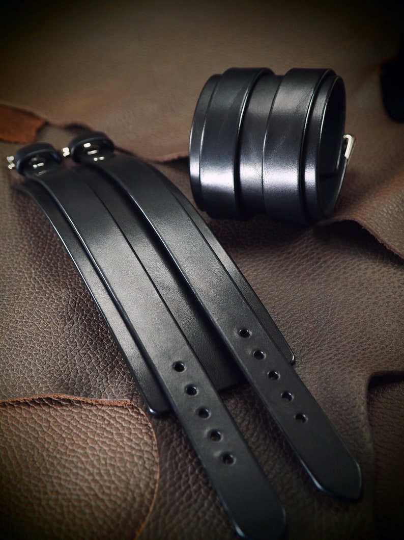 May include: Black leather wrist cuffs and matching bracelets. The cuffs are wide, with multiple stacked bands and a silver buckle. The bracelets have adjustable straps with multiple holes and silver hardware. The items are displayed on a textured brown surface.