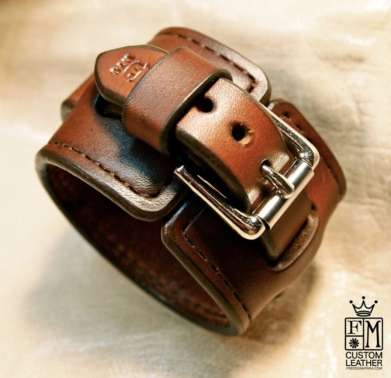 May include: A brown leather cuff bracelet with a silver buckle. The bracelet is stitched with brown thread and has a small logo on the top.