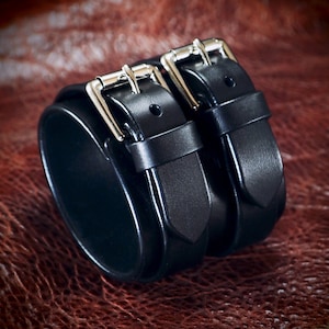 May include: A black leather wrist cuff with two silver buckles. The cuff is thick and wraps around the wrist, with two straps and buckles for closure. The leather has a smooth, polished finish.