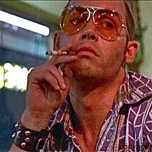 May include: A man wearing a purple and white patterned shirt, a black studded bracelet, and large orange sunglasses is smoking a cigarette. He has a serious expression on his face.