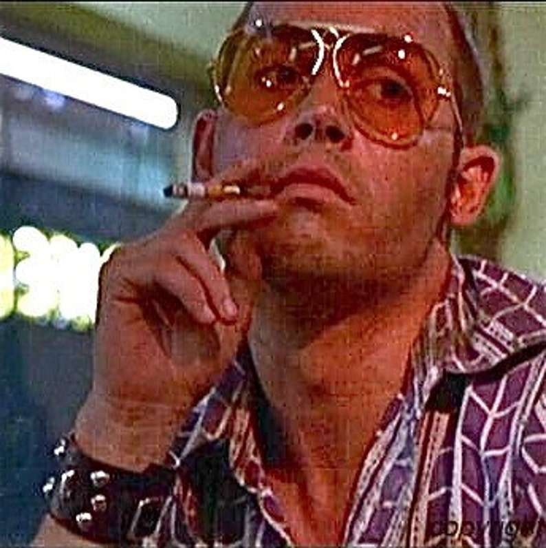 May include: A man wearing a purple and white patterned shirt, a black studded bracelet, and large orange sunglasses is smoking a cigarette. He has a serious expression on his face.