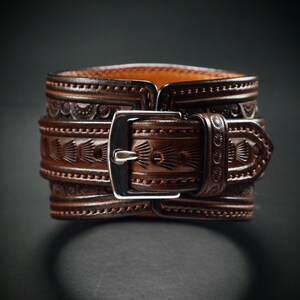 May include: A brown leather cuff bracelet with intricate embossed patterns and a silver-colored buckle. The cuff features detailed designs and is secured with a buckle closure. The leather has a rich, warm tone, and the design is visible on the cuff.