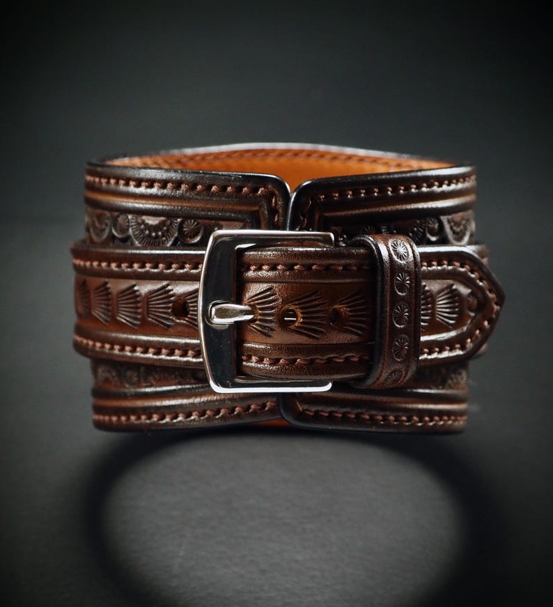 May include: A brown leather cuff bracelet with intricate embossed patterns and a silver-colored buckle. The cuff features detailed designs and is secured with a buckle closure. The leather has a rich, warm tone, and the design is visible on the cuff.