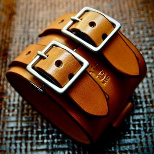Könnte beinhalten: Ein braunes Lederarmband mit doppelter Schnalle und silbernen Schnallen. Das Armband trägt den Schriftzug "NYC".