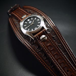 May include: A brown leather watch with a silver watch face and a black dial. The watch is set on a wide, intricately carved leather cuff. The cuff features detailed embossed patterns and is secured with metal rivets.