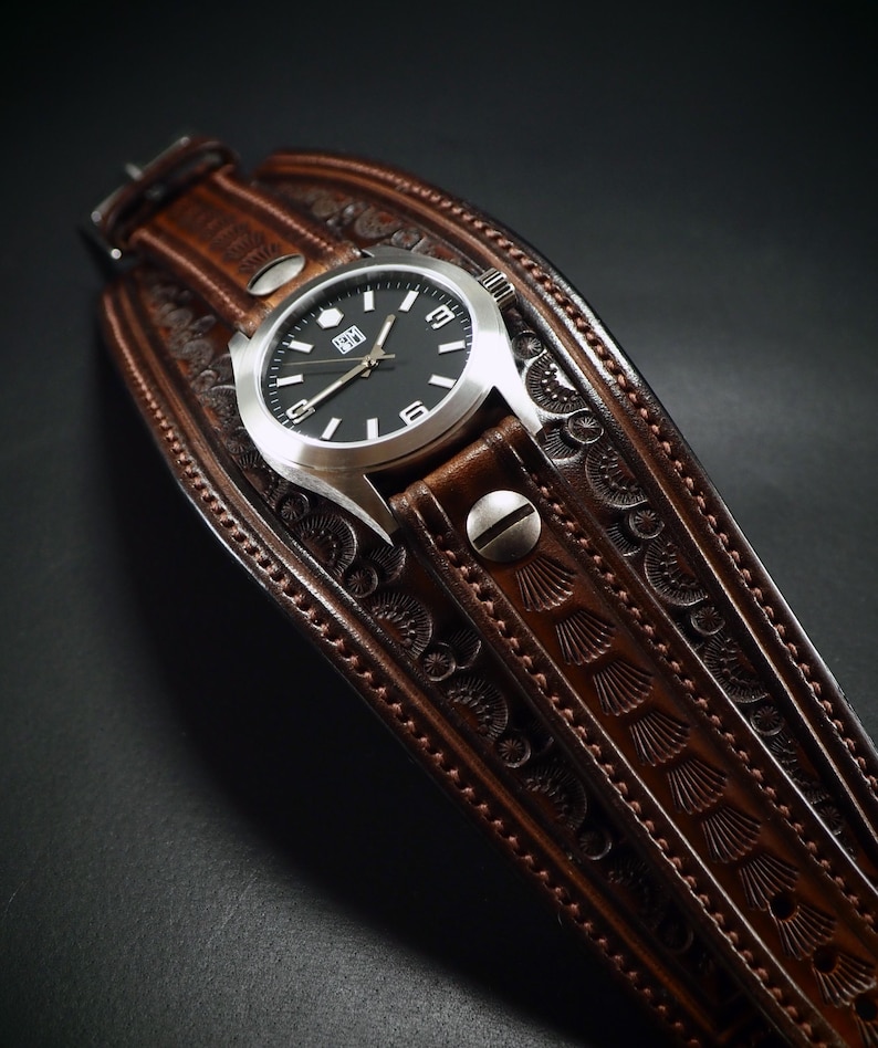 May include: A brown leather watch with a silver watch face and a black dial. The watch is set on a wide, intricately carved leather cuff. The cuff features detailed embossed patterns and is secured with metal rivets.
