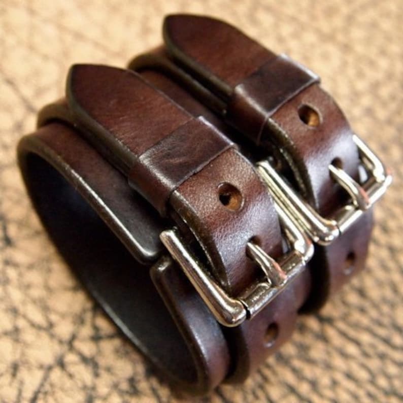May include: A brown leather bracelet with two silver buckles. The bracelet is made of three layers of leather.