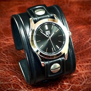 May include: A wristwatch with a round black dial, silver-tone hands, and indices, featuring a logo with the letters RM. The watch is mounted on a wide black leather cuff strap secured with two prominent silver-tone metal rivets at the top and bottom.