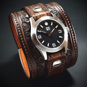 May include: A brown leather cuff watch with a silver watch face and a black dial. The watch has white numerals and hands. The leather cuff is detailed with embossed patterns and metal rivets.