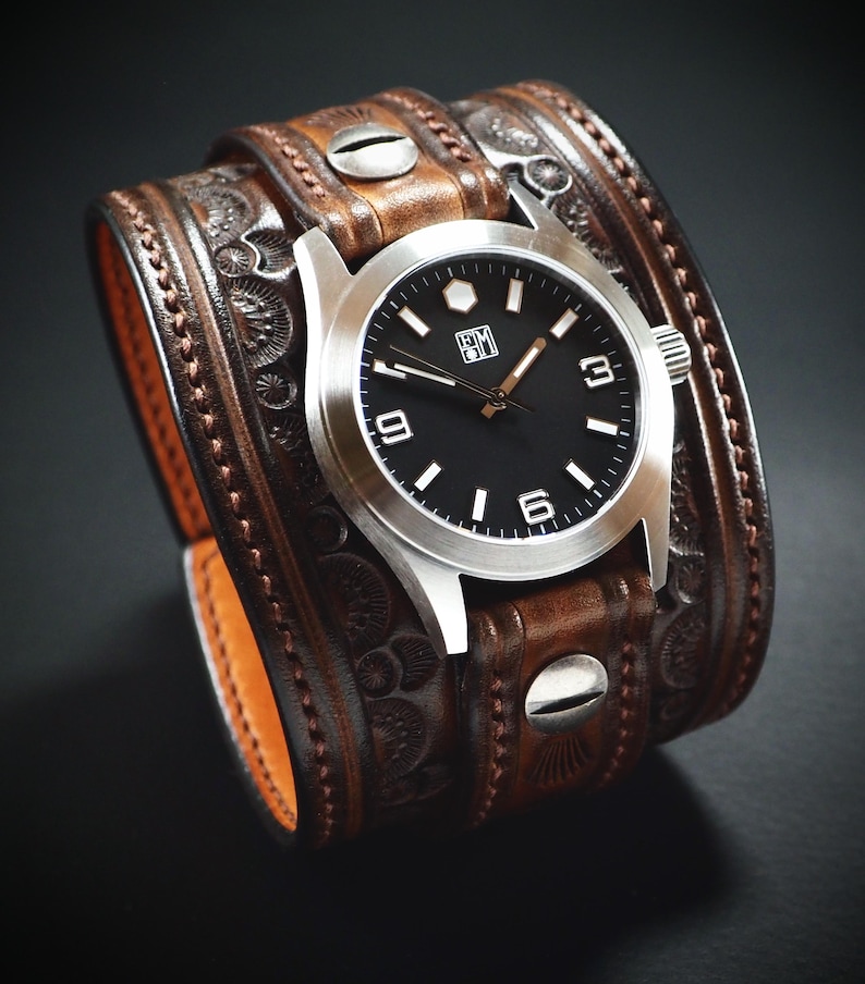 May include: A brown leather cuff watch with a silver watch face and a black dial. The watch has white numerals and hands. The leather cuff is detailed with embossed patterns and metal rivets.