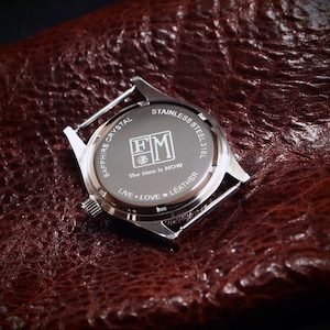 May include: A stainless steel watch back with the text "SAPPHIRE CRYSTAL" and "STAINLESS STEEL 316L". The watch back also has the text "The time is NOW" and "LIVE &bull; LOVE &bull; LEATHER". The watch is on a brown leather surface.