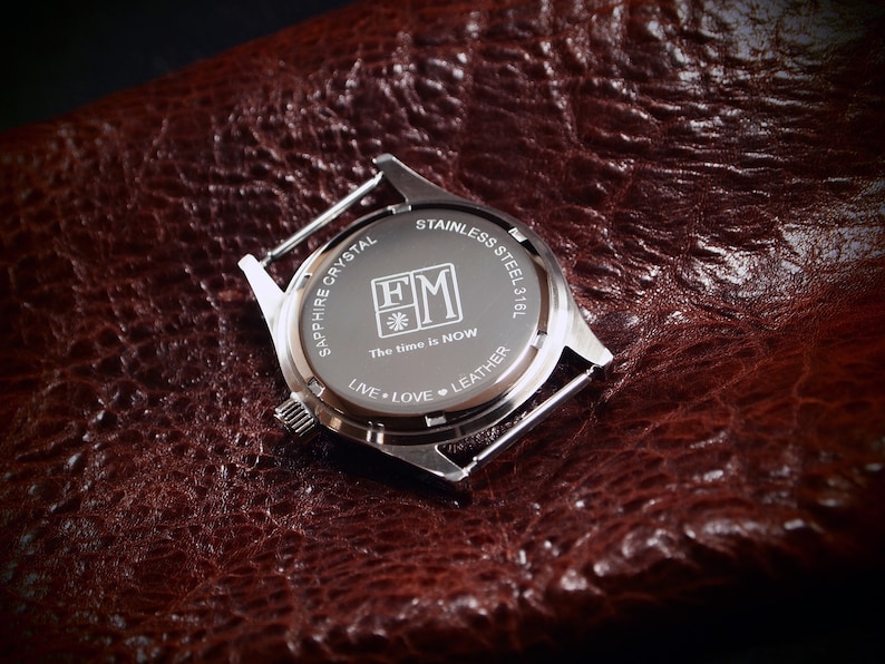 May include: A stainless steel watch back with the text "SAPPHIRE CRYSTAL" and "STAINLESS STEEL 316L". The watch back also has the text "The time is NOW" and "LIVE &bull; LOVE &bull; LEATHER". The watch is on a brown leather surface.