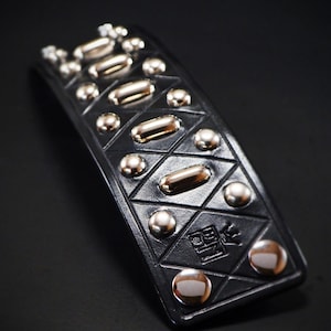 May include: Black leather bracelet with silver and gold-toned studs. The bracelet features a geometric diamond pattern and a small embossed design. The studs are various shapes and sizes, adding visual interest to the accessory.