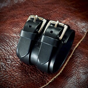 May include: A black leather wrist cuff with two silver buckles. The cuff is made of thick, sturdy leather and features a double-strap design. The buckles are shiny and add a touch of metallic detail to the accessory. The cuff is shown on a brown leather surface.