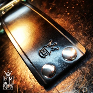 May include: Close-up of a black leather strap with silver metal rivets and a logo that says "FM Custom Leather" with a crown above the letters.